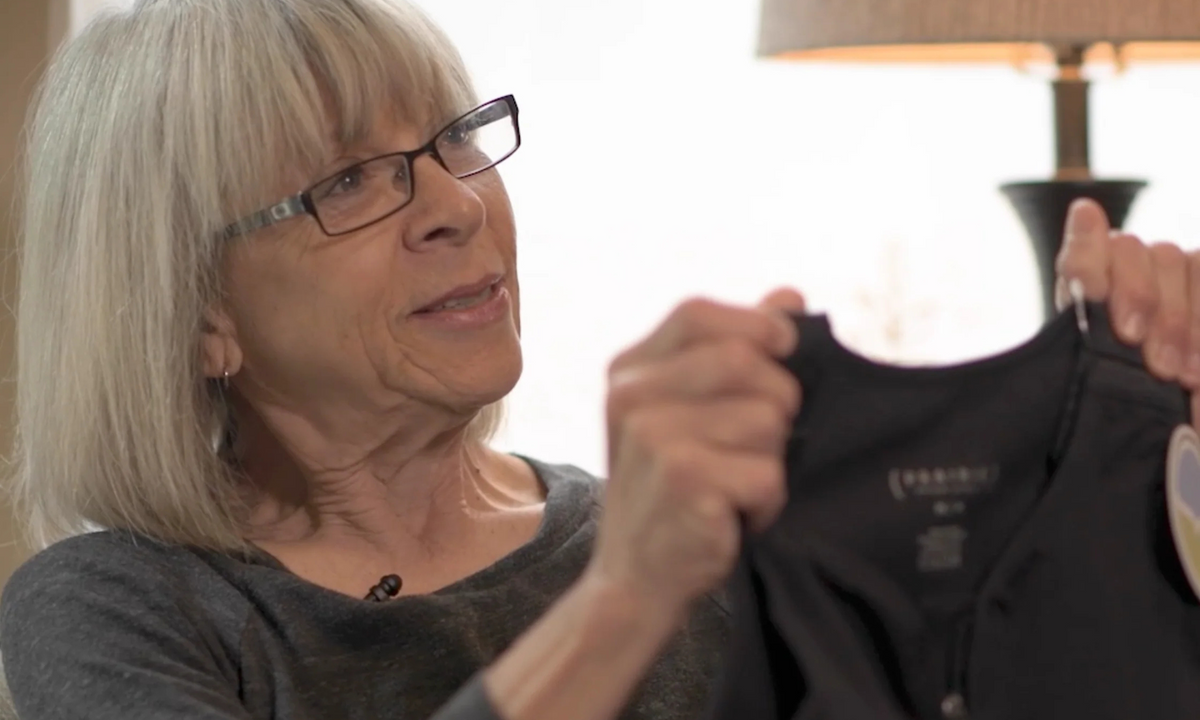 Woman with grey hair and glasses holds up a black Prairie Wear bra with its swing tag while talking