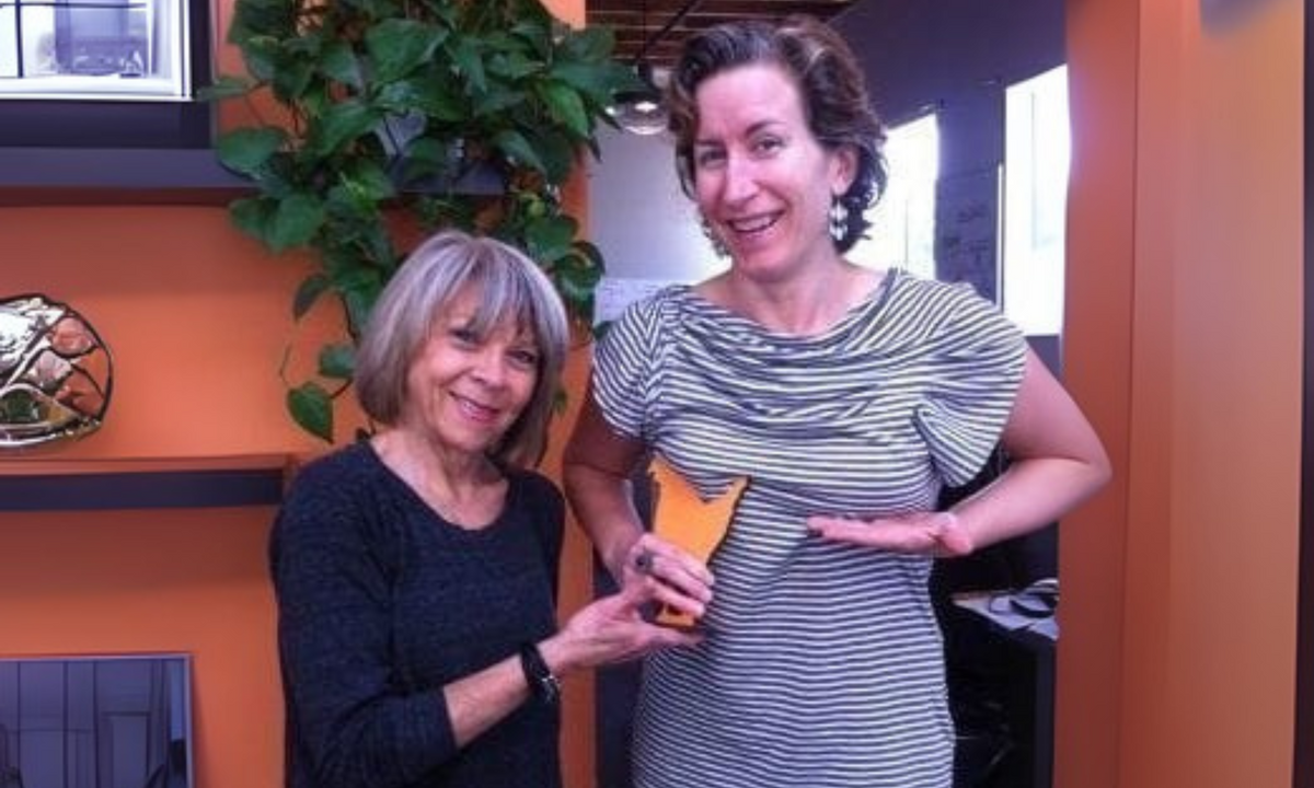 Two smiling women in an office with orange walls hold up a small orange object between them