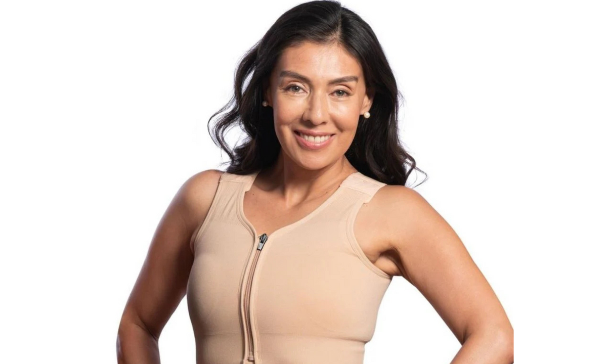 Smiling woman with long dark hair in a beige front-zip compression bra, hands on hips, on a white background