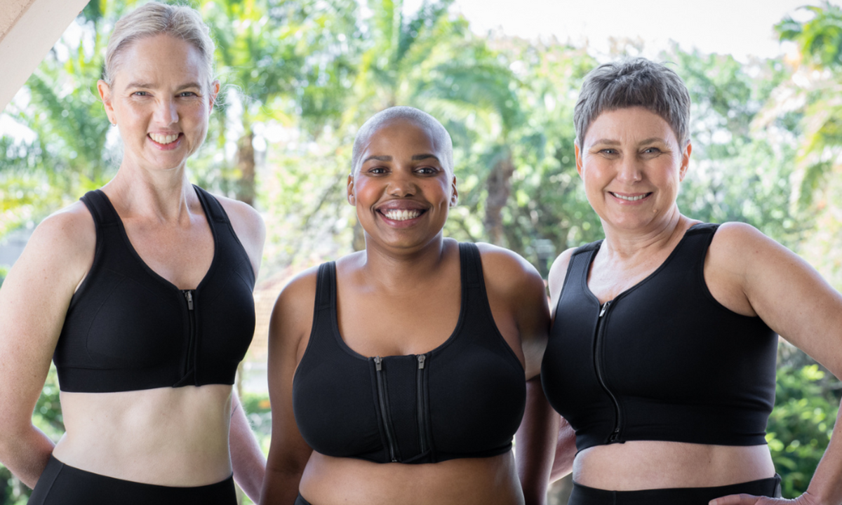 Three smiling women in black front-zip compression bras stand together outdoors with palm trees behind