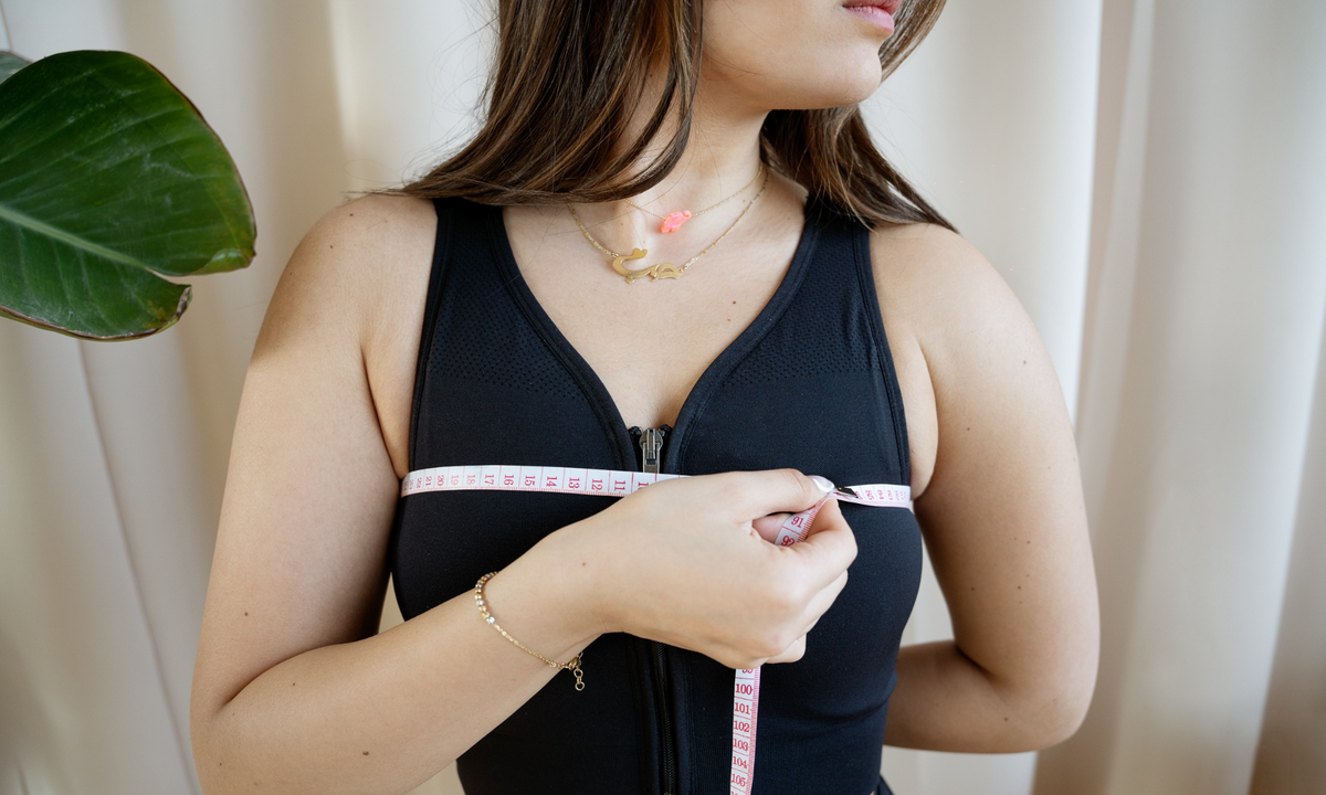 Woman measures her upper chest with a tape measure worn over a black front-zip compression bra