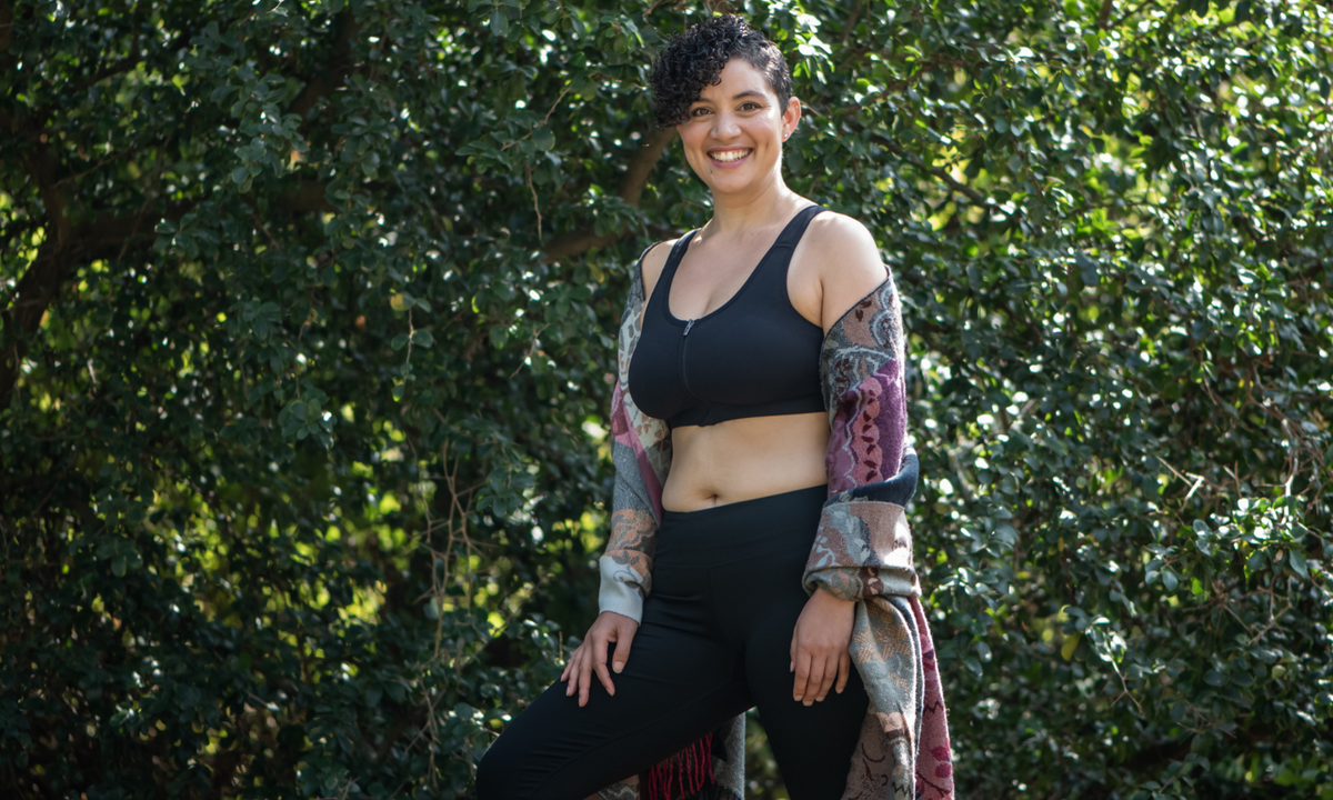 Smiling woman in a black front-zip bra, black leggings and a patterned shawl stands in front of green foliage