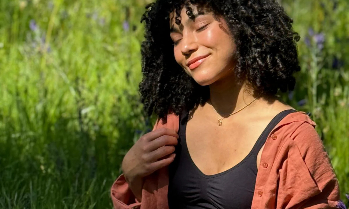 Woman with curly hair smiles, eyes closed, in a sunny meadow in a black everyday bra under an open rust shirt
