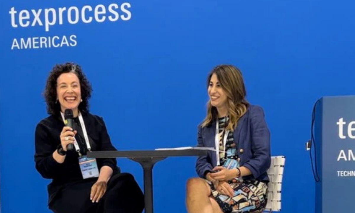 Two women talk on stage at Texprocess Americas, one laughing into a microphone, against a blue backdrop